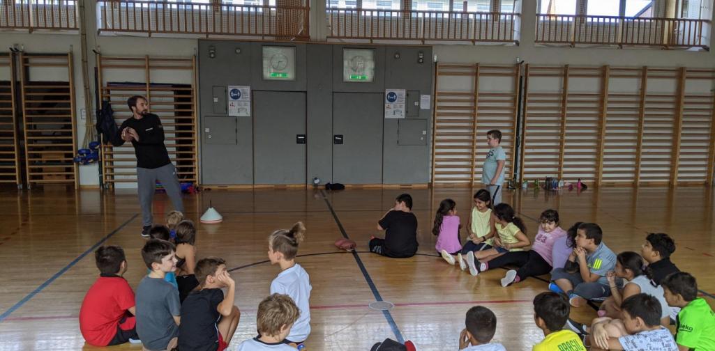 Gruppenbild Flag Football 3a Klasse im Sitzkreis im Turnsaal mit Coach Phil