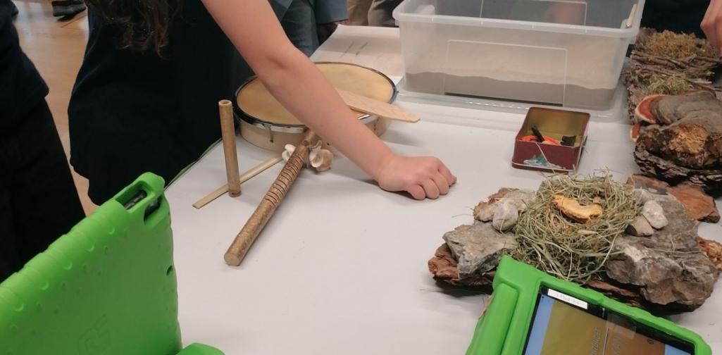 Man sieht Tablets, die auf einem Tisch liegen, zudem sieht man den Arm eines Kindes, welcher gerade auf ein Strohnest hinweist, in Hintergrund sieht man eine Plastikbox, welche mit Sand gefüllt ist